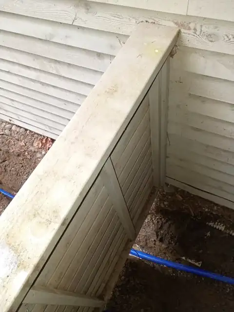 Blue PEX service line routed beneath a wooden utility enclosure against the house siding