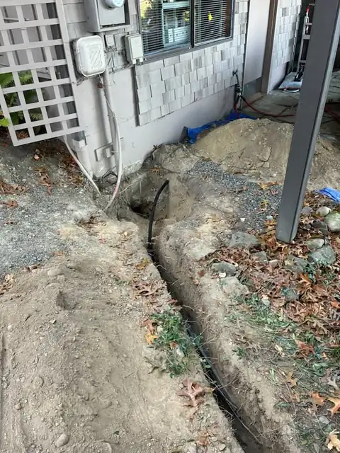Black poly water line laid in a trench along a shingle-sided home with spoils on a tarp