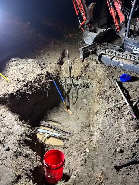 Excavation pit with old pipes exposed at night beside a mini excavator