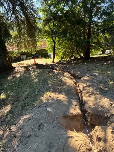 Trench running through a wooded yard toward a home under evergreen trees
