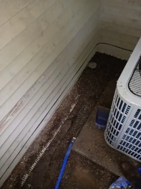 Narrow trench opened along a house wall beside an outdoor AC condenser to reach a leaking supply line