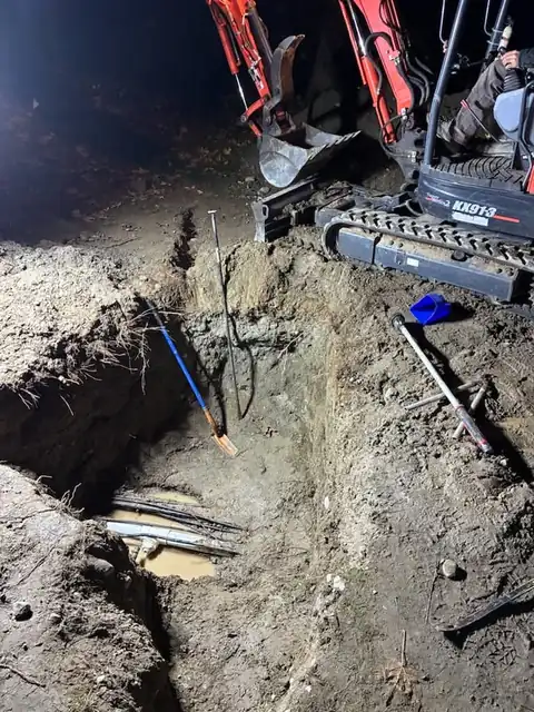 Excavation pit with exposed pipes lit by work lights beside a mini excavator at night