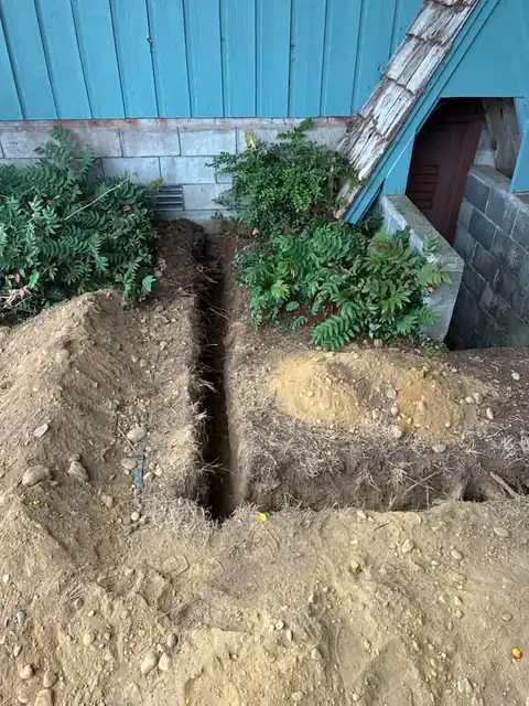Trench dug to a block foundation beside a teal house to reach the water service entry