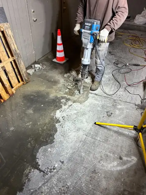 Technician breaking concrete with a demolition hammer to expose a leaking pipe under a slab