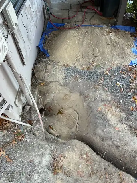 Excavation pit at a house foundation with spoils on a blue tarp and a hose line nearby