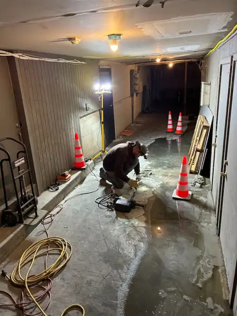 Technician kneeling at an opened slab section in a commercial basement with work lights and cones