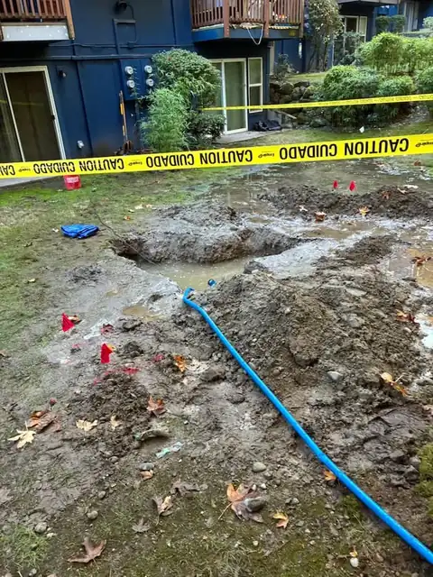 Flooded lawn and excavation at a blue multi-family building with caution tape and a discharge hose