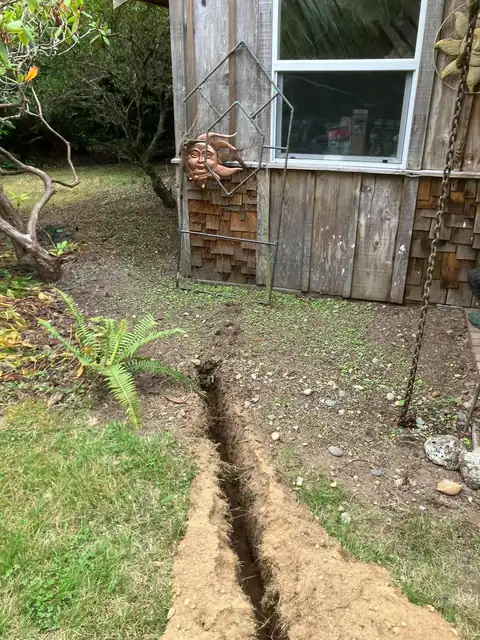Hand-dug trench leading to a cedar-shingled cabin wall