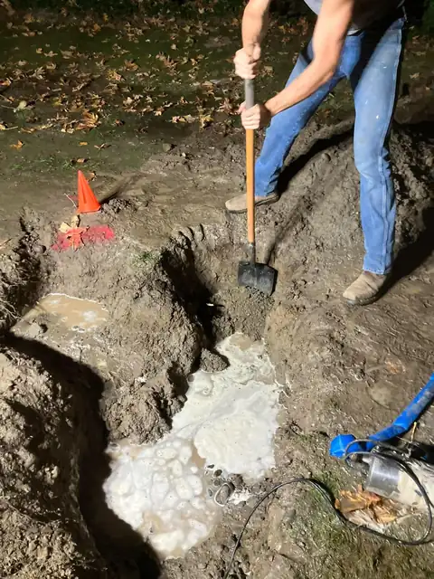Technician shoveling in a muddy, water-filled excavation pit at night