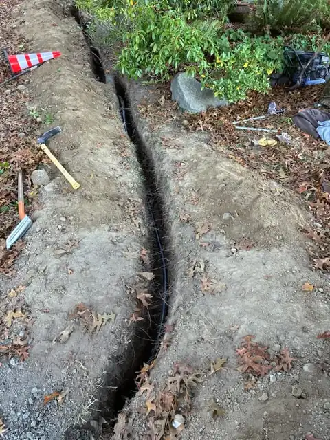 Black poly water line in a trench with hand tools and a cone on a wooded lot