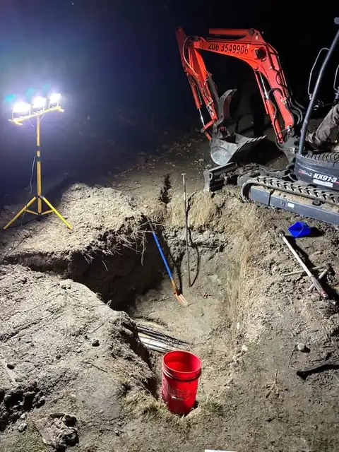 360 Leak Finders mini excavator with the company phone number on the boom beside a night excavation