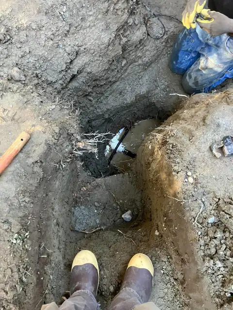 Pipe fitting and leak exposed at the bottom of a hand-dug pit with a technician kneeling nearby