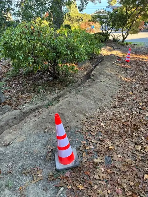 Safety cone marking a backfilled trench beside shrubs on a residential lot