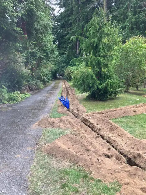 Long trench along a paved driveway with a coil of blue water line standing ready
